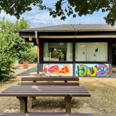 Sitzgruppe mit Holzbänken und Tisch im Außenbereich vor einem Gebäude mit bunten Graffiti.