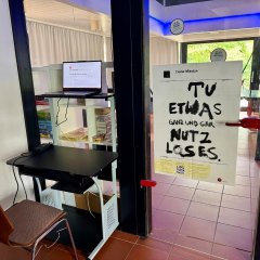 Eingangsbereich mit Computerarbeitsplatz und einem Plakat an der Glastür mit der Aufschrift „Tu etwas ganz und gar Nutzloses“.