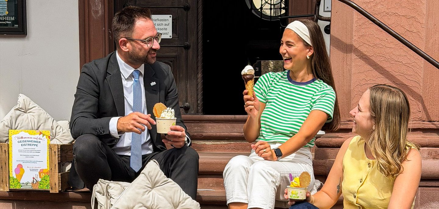 Bürgermeister Christian Aßmann und Mitarbeitende der Stadtverwaltung essen gemeinsam Eis vom Eiscafé Pilot auf der Treppe des Rathausgebäudes.
